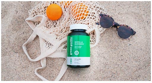 A mesh bag of oranges and sunglasses on a beach next to a jar of Green Gorilla™ gummies.