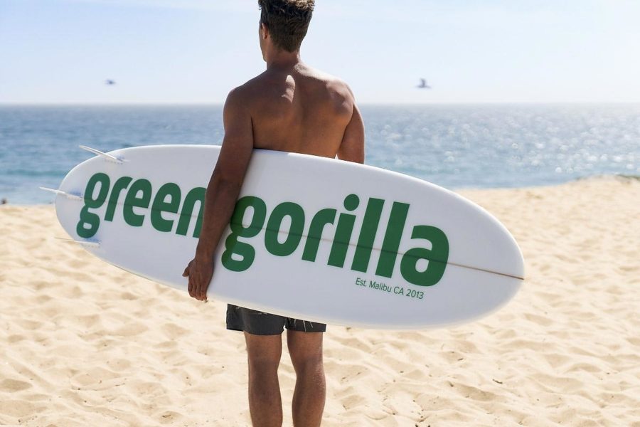 Man in swimsuit holding a surfboard that says “Green Gorilla™.”
