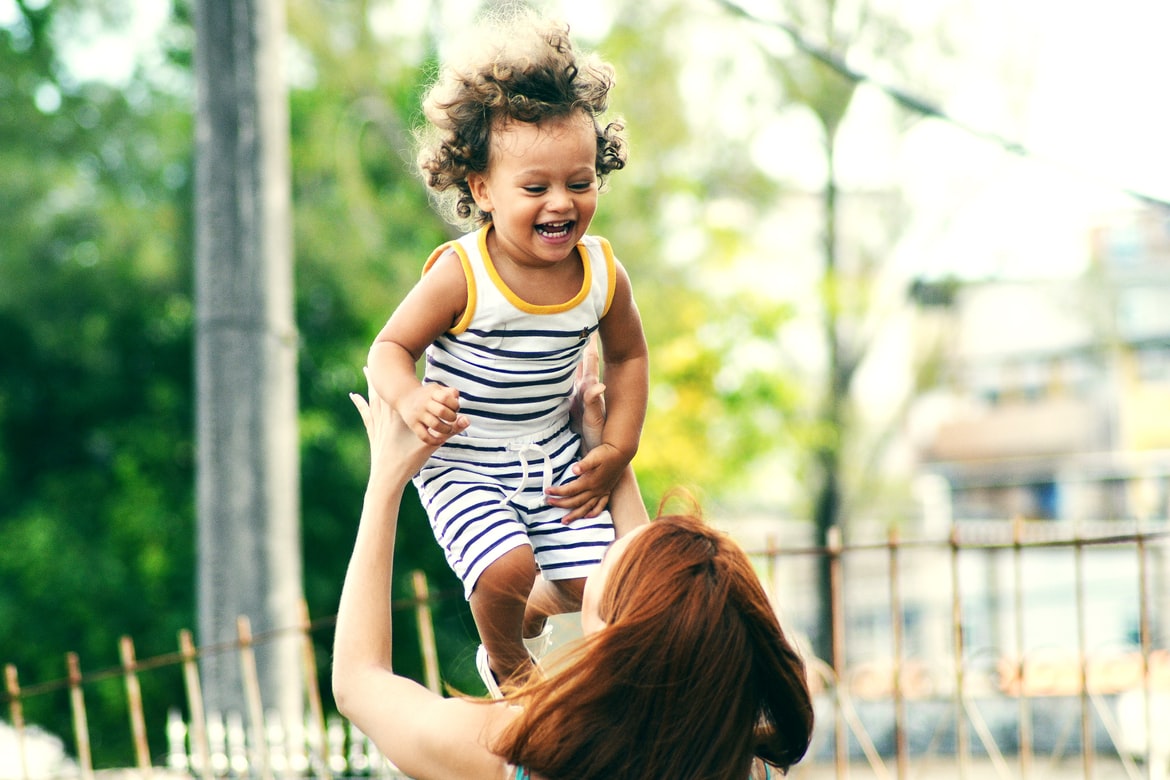 Woman tossing a smiling toddler with a striped outfit and curly hair into the air