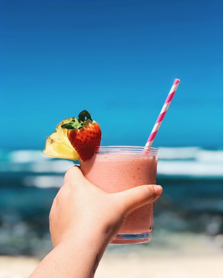 A person holding a smoothie with a pineapple chunk and strawberry on the rim